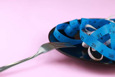 Pembe arka planda izole edilmiş siyah seramik plaka üzerindeki mavi mezura ve çatal. Kilo verme kavramı için diyet.