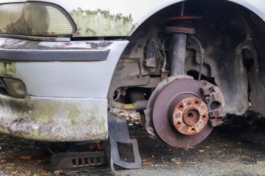 Terk edilmiş eski bir arabanın ön tekerlek kuyusuna yakın çekim. Tekerlek sökülmüş, paslı fren diski ve kalibre. Aracın vücudu kir ve yosunla kaplı..