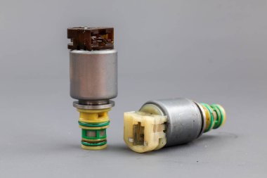 İki Allison iletim solenoid vanası gri bir arka planda gösteriliyor, biri dik duruyor ve diğeri yan tarafında. Fotoğraf metal gövdelerini, kablolarını ve konnektörlerini vurguluyor..