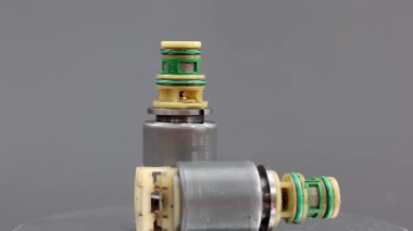 İki Allison iletim solenoid vanası gri bir arka planda gösteriliyor, biri dik duruyor ve diğeri yan tarafında. Fotoğraf metal gövdelerini, kablolarını ve konnektörlerini vurguluyor..