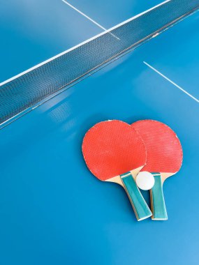 İki kırmızı masa tenisi raketi ve ağların yanındaki mavi masa tenisi masasında dinlenen beyaz bir top. Spor ve eğlence için dinamik bir sahne.