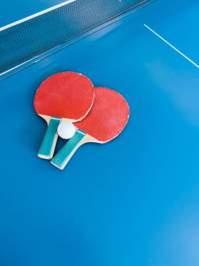 Bir çift kırmızı masa tenisi raketi ve ağların yanındaki mavi masa tenisi masasında dinlenen beyaz bir top. Rekabetçi spor ve eğlenceyi temsil eden bir sahne.
