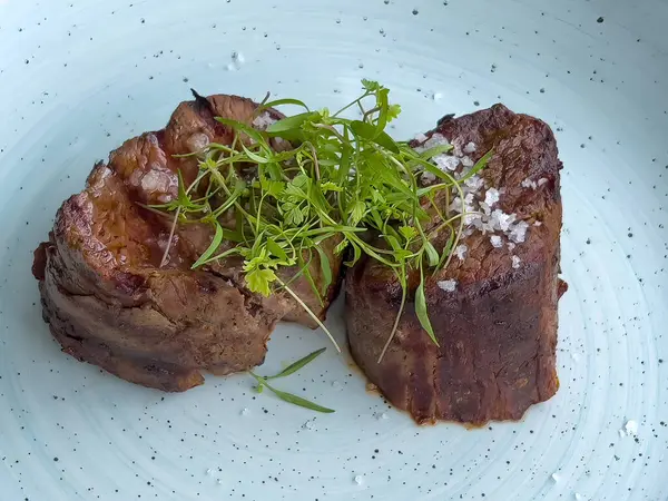 Deniz tuzu ve taze otlarla kaplı sulu ızgara fileminyon biftek. Mavi seramik tabakta servis ediliyor. Zengin aromalı ve dokulu bir gurme et yemeği..