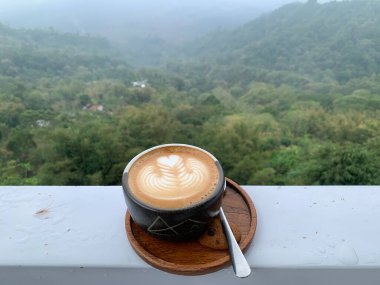 Çiselemeden sonra balkondaki kahverengi ahşap çay tabağına izole edilmiş lattee resimli bir fincan kahve. Fotokopi alanı, web siteleri, tasarım veya duvar kağıdı için arka plan olarak Bokeh sisli ve puslu yeşil doğa.