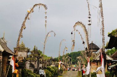 Penjor bambu kutupları Endonezya 'nın Bali kentindeki Penglipuran Köyü' nün taş sokağında dramatik bulutların altında. Geniş açılı. Seyahat, kültür, maneviyat ve sokak hayatı temalarına uyuyor. 21 Kasım 2024.