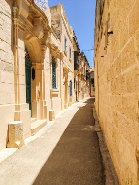 Malta 'da güneşli bir yaz gününde, sıcak güneş ışığıyla yıkanmış boş bir sokak. Sessiz, kaldırım taşı caddesi geleneksel Malta mimarisi ile kaplı. Adanın cazibesini vurgulayan huzurlu ve resimli bir manzara yaratıyor..