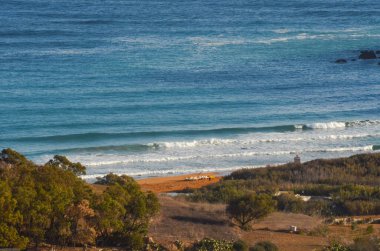 Yumuşak okyanus dalgalarının kumlu sahilde yuvarlandığı pitoresk bir kıyı manzarası. Bereketli yeşil ağaçlarla çevrili bu sakin manzara, sakin mavi sularla doğanın güzelliğini yakalar..