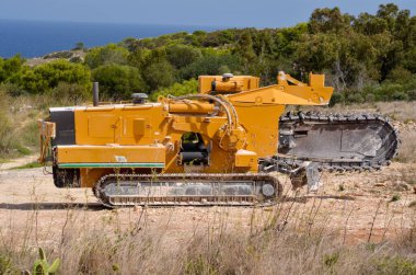Sahilde manzaralı bir arka planda endüstriyel siper kazma makinesi çalışıyor. Parlak sarıya boyanmış bu güçlü inşaat ekipmanları engebeli arazilerde verimli siper kazma görevleri için tasarlanmıştır..