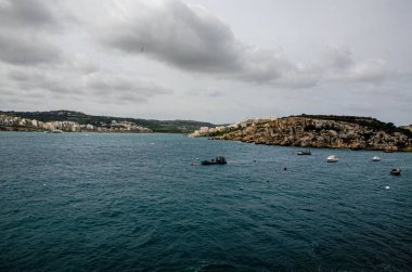 Malta, Mellieha 'nın kayalık kıyı şeridinde yüzen balıkçı tekneleri ve eğlence gemileri. Kentsel geçmişe sahip sakin bir deniz manzarası