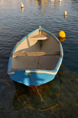 Rıhtıma bağlı yıpranmış beyaz bir kayık, sakin liman sularında huzur içinde yüzer. Gün batımının hafif yansımaları yüzeye vurur.. 