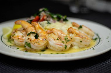 Sarımsaklı tereyağı sosunda sallanan, otlarla süslenmiş ve kremalı tabanlı lezzetli karidesler. Beyaz tabakta nefis deniz ürünleri tabağı, restoran menüleri, yemek siteleri ve gurme yemek tasarımı için ideal.