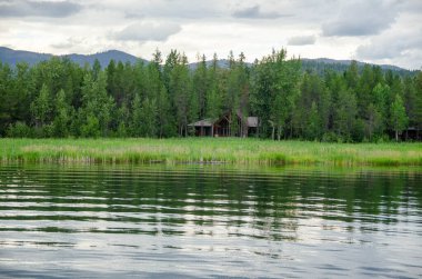 Ağaçların arasına saklanmış sessiz bir orman kulübesi, yumuşak dalgalarla dolu huzurlu bir göl kıyısına bakıyor.