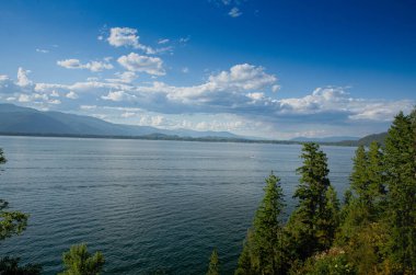 Bereketli yeşillik ve uzak dağlarla çevrili, bulutlarla dolu güzel bir gökyüzünün altında nefes kesici bir göl manzarası.