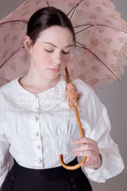 Beyaz keten keten bir bluz ve siyah etek giyen ve elinde klasik bir güneş şemsiyesi tutan Viktorya Dönemi 'nden genç bir kadın.