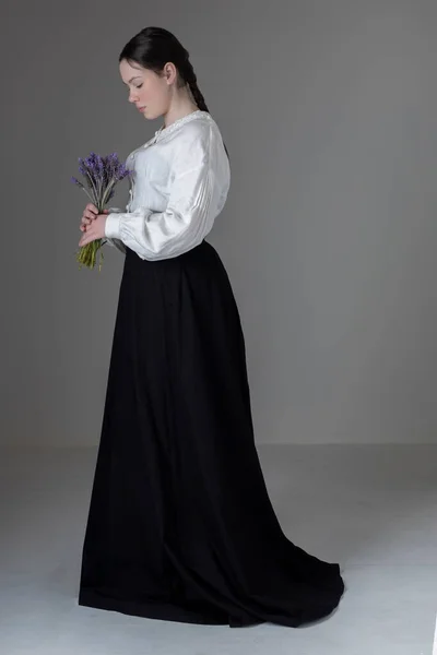 A young Victorian or Edwardian woman wearing a white linen Garibaldi blouse and black skirt and holding a bunch of lavender