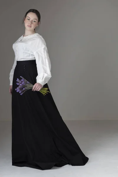 白いリネンのガリバルディのブラウスと黒いスカートを着てラベンダーの束を保持している若いビクトリア朝またはエドワード朝の女性 ロイヤリティフリーのストック写真