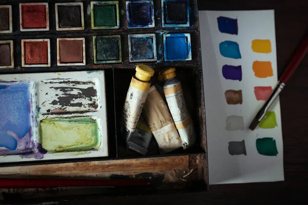 A vintage water color set in a metal tin sitting on a dark wood desk with color swatches on watercolor paper