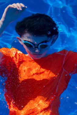 Gözlük takan ve portakallı yüzme bluzu giyen ve suyun altında kabarcıklar çıkaran bir çocuk.