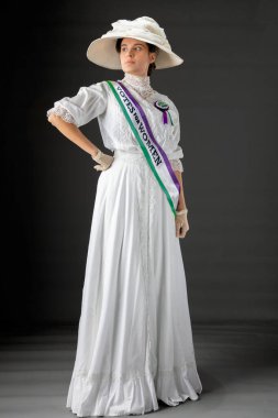 Birleşik Krallık Viktorya dönemi ya da Edwardian Suffragette 'i tarihsel olarak doğru mor, yeşil kuşak ve roset takıyor ve bir stüdyo arkaplanına karşı kadınların oy kullanma haklarını protesto ediyor.