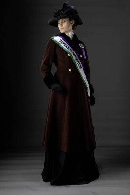 Birleşik Krallık Viktorya dönemi ya da Edwardian Suffragette 'i tarihsel olarak doğru mor, yeşil kuşak ve roset takıyor ve bir stüdyo arkaplanına karşı kadınların oy kullanma haklarını protesto ediyor.