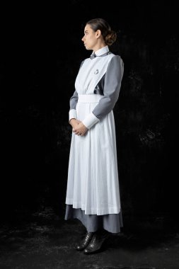 Birinci Dünya Savaşı hemşiresi gri elbiseli ve beyaz önlüklü siyah bir fonda duruyor.