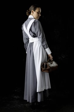 Birinci Dünya Savaşı hemşiresi gri elbiseli ve beyaz önlüklü siyah bir fonda duruyor.