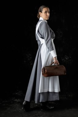 Birinci Dünya Savaşı hemşiresi gri elbiseli ve beyaz önlüklü siyah bir fonda duruyor.