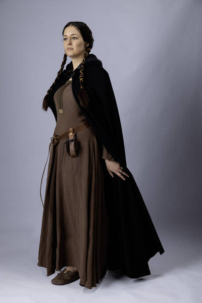 A Medieval woman wearing a kirtle made from rough brown linen fabric. She has long dark hair tied in two long plaits and is standing against a plain backdrop
