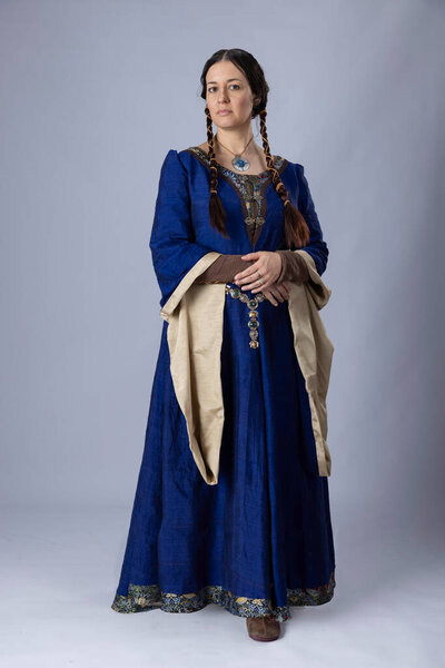 Средневековая женщина в синем шелковом платье. У нее длинные темные волосы, сплетенные в две косы, и она стоит на обычном фоне.