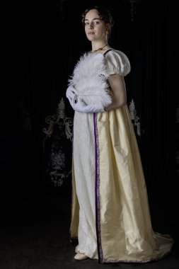 Gece kıyafeti giyen bir naip kadın, nakışlı bir elbise ve uzun bir spencer. Elinde büyük, beyaz bir devekuşu tüyü yelpazesi var.