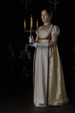 Gece kıyafeti giyen bir naip kadın, nakışlı bir elbise ve uzun bir spencer. Elinde bir şamdan tutuyor.