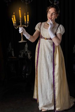 Gece kıyafeti giyen bir naip kadın, nakışlı bir elbise ve uzun bir spencer. Elinde bir şamdan tutuyor.