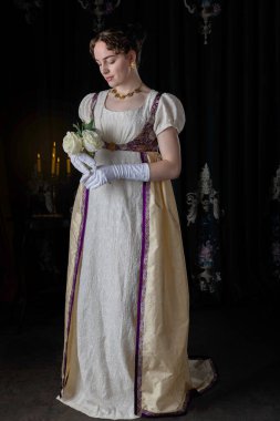 Gece kıyafeti giyen bir naip kadın, nakışlı bir elbise ve uzun bir spencer. Elinde beyaz güller var..