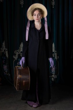 Seyahat kıyafeti giyen bir naip kadın. Ceketi, şapkası ve eldivenleri var. Deri bir şapka kutusu var.