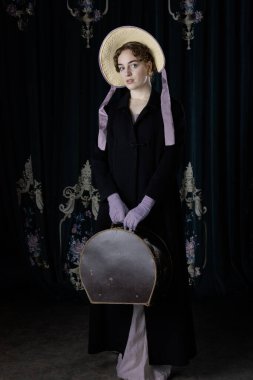 Seyahat kıyafeti giyen bir naip kadın. Ceketi, şapkası ve eldivenleri var. Deri bir şapka kutusu var.