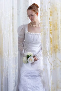 Beyaz pamuklu elbise giyen ve arkadan ışıklandırılmış dantel perdelerle çevrili bir naip kadın.