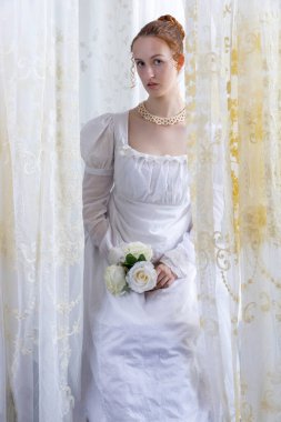 Beyaz pamuklu elbise giyen ve arkadan ışıklandırılmış dantel perdelerle çevrili bir naip kadın.