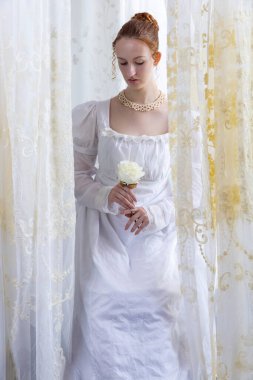 Beyaz pamuklu elbise giyen ve arkadan ışıklandırılmış dantel perdelerle çevrili bir naip kadın.