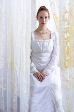 Beyaz pamuklu elbise giyen ve arkadan ışıklandırılmış dantel perdelerle çevrili bir naip kadın.
