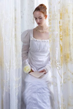 Beyaz pamuklu elbise giyen ve arkadan ışıklandırılmış dantel perdelerle çevrili bir naip kadın.