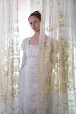 Beyaz pamuklu elbise giyen ve arkadan ışıklandırılmış dantel perdelerle çevrili bir naip kadın.