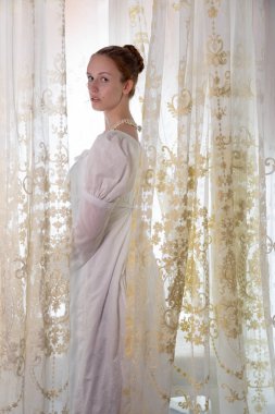 Beyaz pamuklu elbise giyen ve arkadan ışıklandırılmış dantel perdelerle çevrili bir naip kadın.