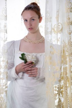 Beyaz pamuklu elbise giyen ve arkadan ışıklandırılmış dantel perdelerle çevrili bir naip kadın.