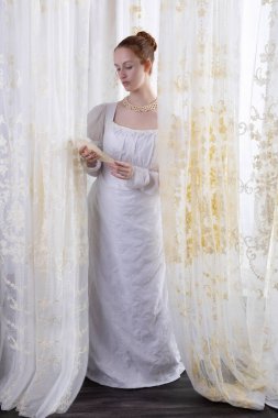 Beyaz pamuklu elbise giyen ve arkadan ışıklandırılmış dantel perdelerle çevrili bir naip kadın.