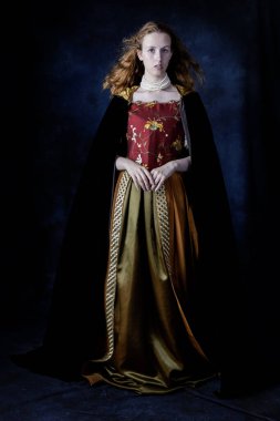 Rönesans kadını, kızıl saçlı, işlemeli ipek bir elbise giyen stüdyo arkaplanına yaslanmış.