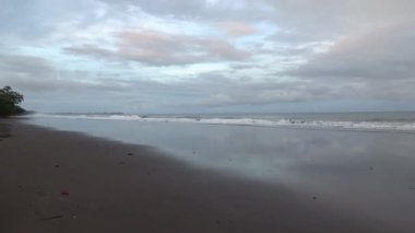 Sakin dalgalarla sakin bir sahil sahnesi ve ıslak kuma yansıyan bulutlu bir gökyüzü..