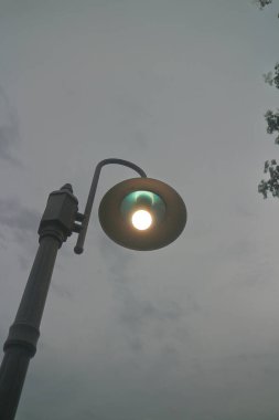 Bulutlu bir gökyüzünde parlayan yuvarlak ışıklı bir sokak lambası. Lamba uzun bir direğe monte edilmiş, modern bir tasarım sergiliyor..