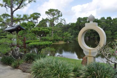 A view of the lake at Morikami Japanese Botanical Gardens in Boynton Beach Florida with landscaping, sculpture and boulders in the foreground on a sunny summer day with blue skies and clouds clipart