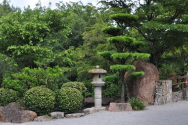 Boynton Beach Florida 'daki Morikami Japon Botanik Bahçeleri' nin yürüme yolundan gelen çalılar, heykeller ve kayalarla yapılan manzara. Güneşli bir yaz gününde arka planda mavi gökyüzü ve bulutlarla kaplı ağaçlar.