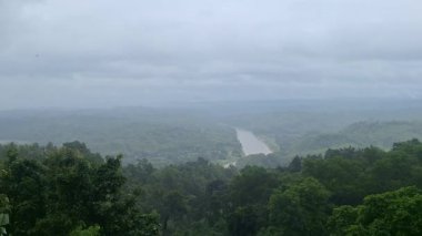 Sakin bir nehir bulutlu bir gökyüzünün altındaki yemyeşil bir ormanın içinden akar ve huzurlu bir doğal görüntü oluşturur.